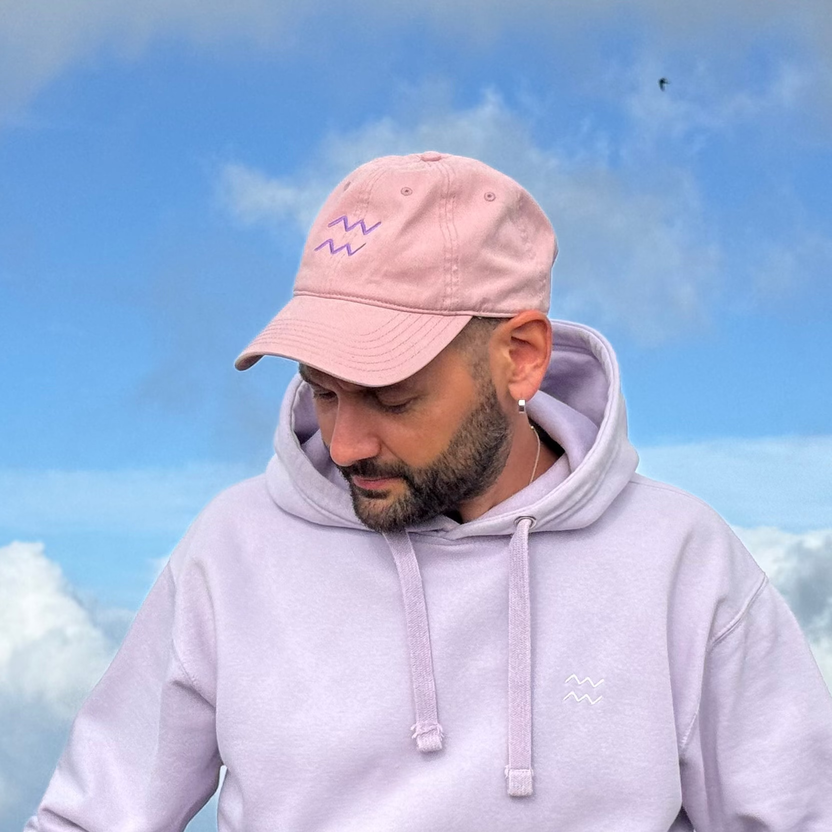 A man wearing a light pink baseball cap and a pale lavender hoodie, sitting outdoors against a dramatic blue sky with clouds.