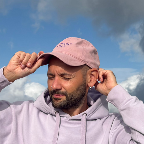A man wearing a lavender hoodie and matching pink cap stands outdoors against a dramatic blue and gray sky with mountains in the background, adjusting his cap.