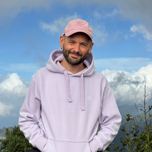 A man wearing a light lavender hoodie and a pink cap stands outdoors against a dramatic blue and white sky background.