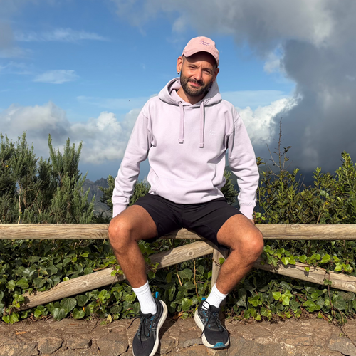 A man sitting on a wooden fence outdoors, wearing a light pink hoodie, black shorts, a pink cap, white socks, and black running shoes, with green foliage and a cloudy blue sky behind him.