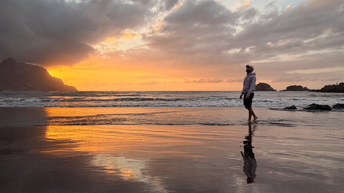 A man stands in shallow ocean water on a beach at sunset, with dramatic clouds, golden light, dark rocks, and a mirror-like reflection on the wet sand.