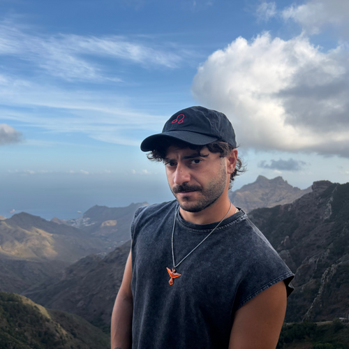 Black zodiac baseball cap of Gamla Healing worn by a man wearing a dark gray sleeveless t-shirt and a silver necklace with an orange cross pendant, standing in front of a scenic mountain and sea background under a blue sky with clouds.