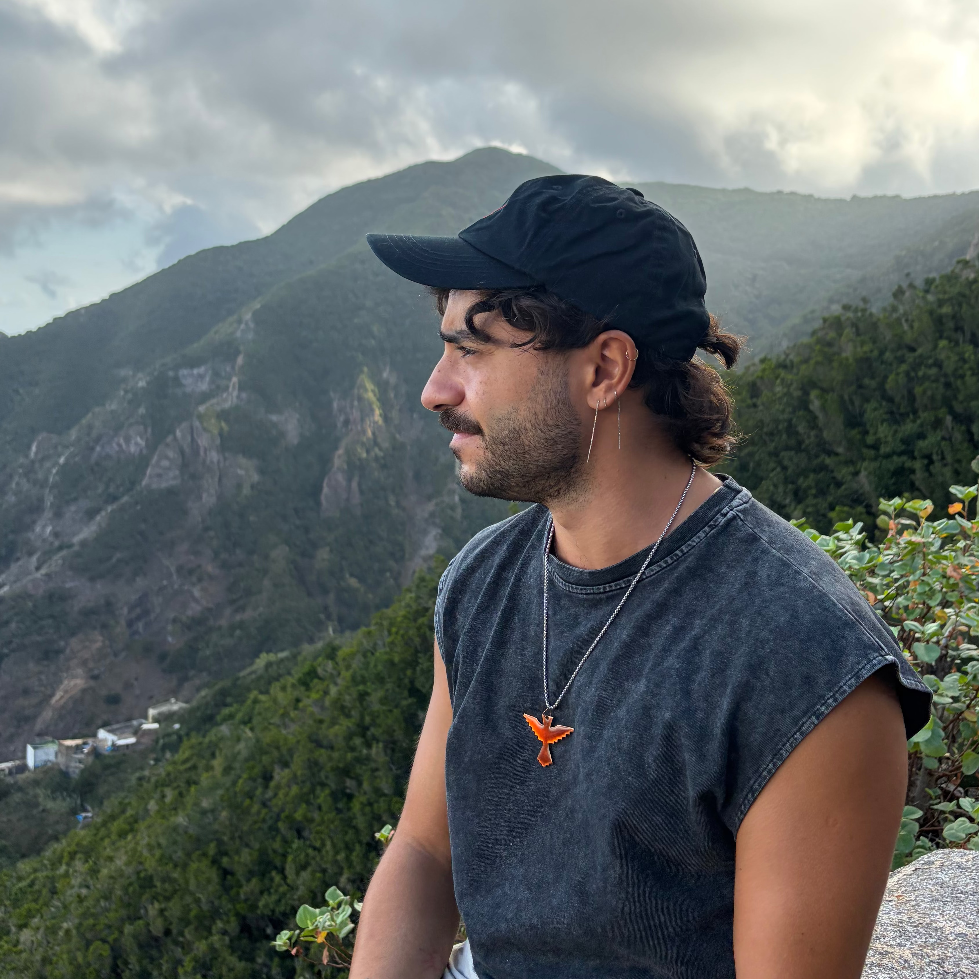Black zodiac baseball cap of Gamla Healing worn by a man wearing a faded black sleeveless t-shirt, and light denim shorts sits outdoors on a stone wall with a mountain landscape and cloudy sky in the background.
